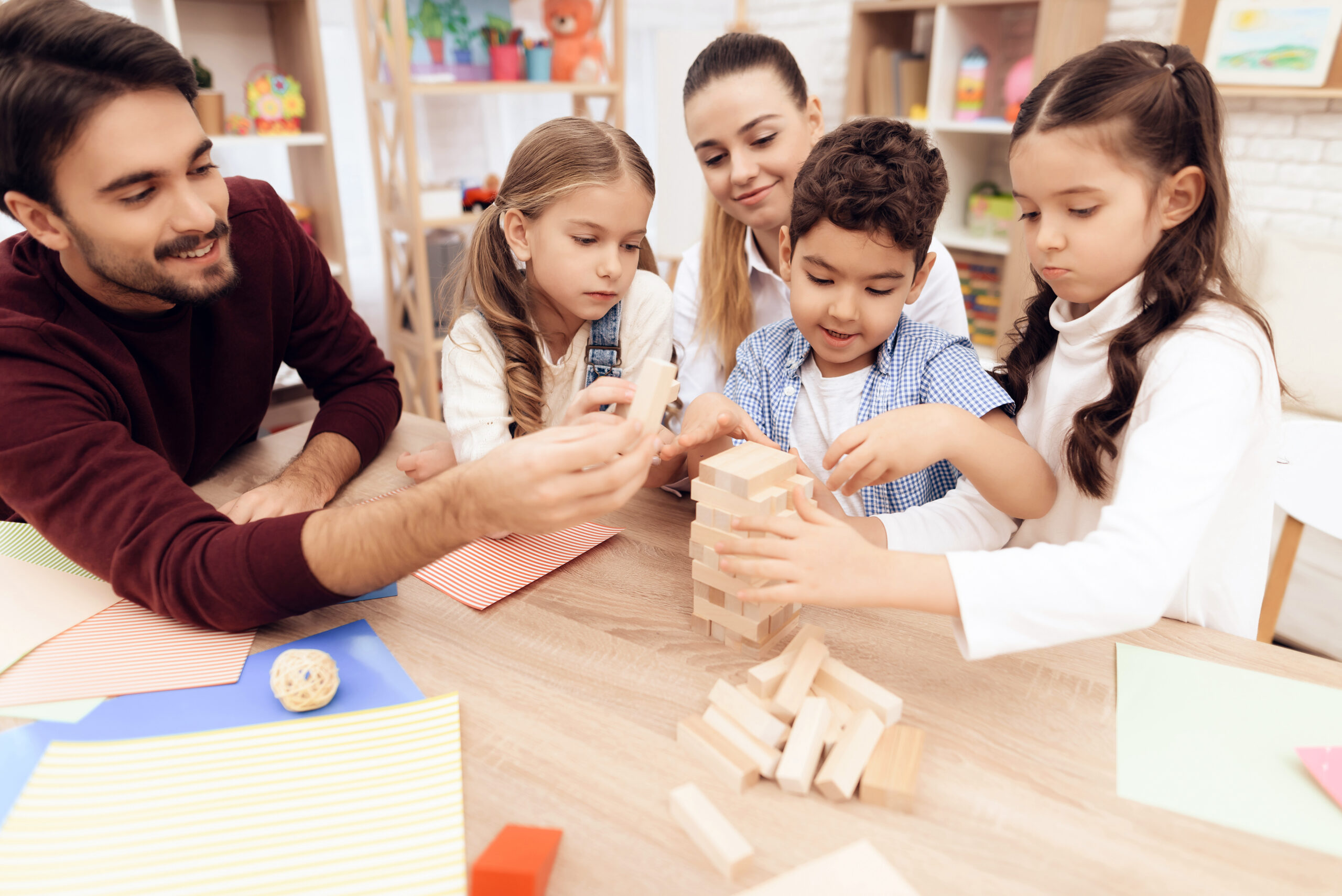 Three young children building a jenga block tower with two adults watching.