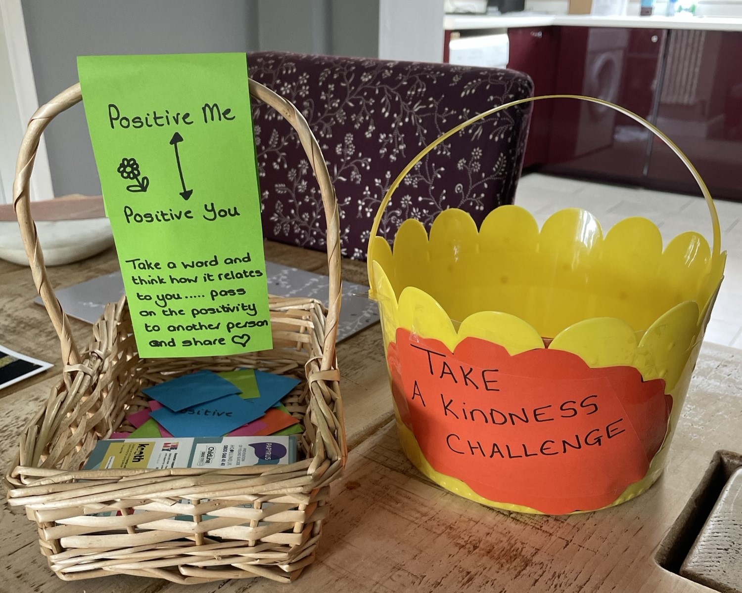 A wicker basket filled with notes and a yellow bucket. The bucket has a label: "Take a kindness challenge". The basket has a message attached: "Positive Me, Positive Y ou. Take a word and think how it relates to you... Pass on the positivity to another person and share"