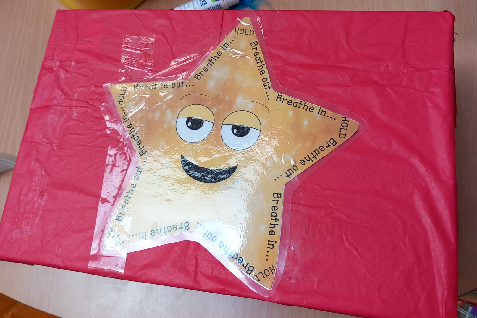 a star with a smiling face, with the words "breathe out...breathe in...HOLD" written around the edges, attached to box surrounded in red crepe paper